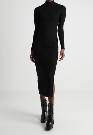 Robe midi noire cintrée en maille côtelée avec manches longues et col montant, associée à des bottines noires brillantes à bout pointu et talon bloc.