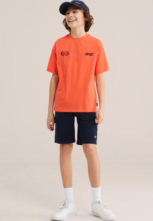Lachende jongen met een marineblauwe pet, oranje T-shirt met print, marineblauwe broek met zakken, witte sokken en witte sneakers tegen een beige achtergrond.