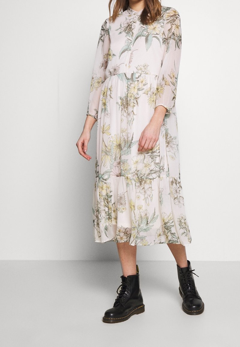 Femme portant une longue robe fleurie claire à manches longues, associée à des bottes de combat noires à lacets, debout devant un fond uni.