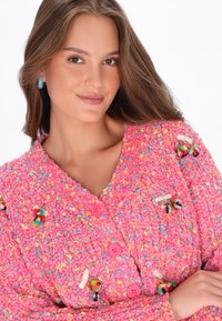 Adīts rozā krekls ar krāsainiem iestrādājumiem, ar lieliem rozā pogām un dekoratīviem pom-pomiem. Mīksta, teksturēta auduma ar ērtu dizainu.