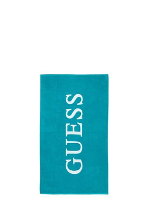 Asciugamano rettangolare color teal con la parola "GUESS" scritta verticalmente in grandi lettere maiuscole bianche.