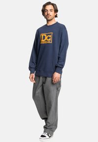 Navy sweatshirt with DC Shoes logo in yellow, round neckline, ribbed cuffs, paired with baggy gray jeans and black-and-white sneakers.