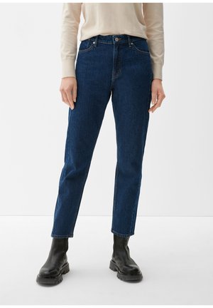 Person iført mørkeblå jeans med lige ben, beige langærmet top og sorte chunky støvler, stående mod en ensfarvet hvid baggrund.