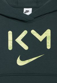Sudadera con capucha de color verde oscuro con patrones abstractos de rayas amarillo-verdosas sobre grandes letras "KCM" y un logotipo de Nike swoosh debajo.