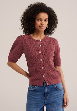 Femme aux cheveux bouclés portant des boucles d'oreilles en anneaux, un pull texturé à manches courtes mauve avec des boutons, et un jean bleu, posant avec la main sur la hanche.