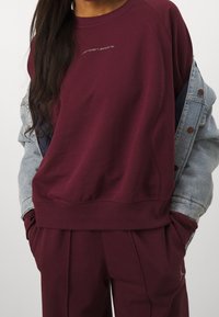 Sudadera burdeos con mangas raglán, que presenta un pequeño logo gris en el pecho. Combinada con pantalones a juego y una chaqueta de mezclilla clara.