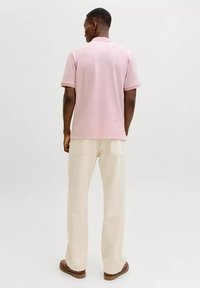 Ein Mann steht mit dem Rücken zur Kamera, trägt ein hellrosa kurzärmeliges Polohemd, cremefarbene Hosen und braune Schuhe vor einem schlichten Hintergrund.