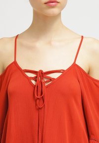 Blouse rouge fluide avec un décolleté en V, devant lacé ajustable et fines bretelles. Présente un tissu texturé et des manches délicatement drapées.