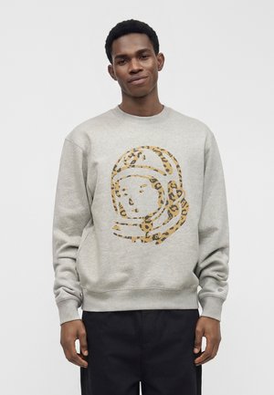 Jeune homme portant un sweat-shirt gris clair avec un grand motif de casque d'astronaute imprimé léopard, debout sur un fond uni.
