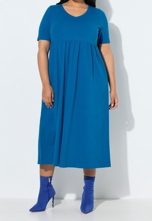 Femme portant une robe bleue longueur genoux avec des manches courtes et des bottines bleues à talons hauts, debout devant un fond blanc.