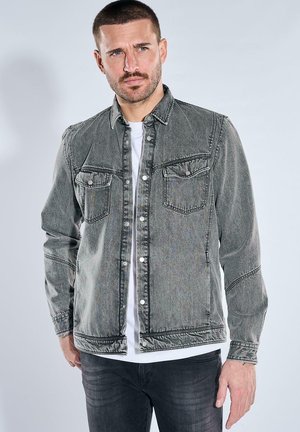 Mann mit kurzen Haaren und Bart, der eine verblasste graue Jeansjacke mit Knöpfen über einem weißen Hemd und dunklen Hosen trägt, vor einfachem Hintergrund.