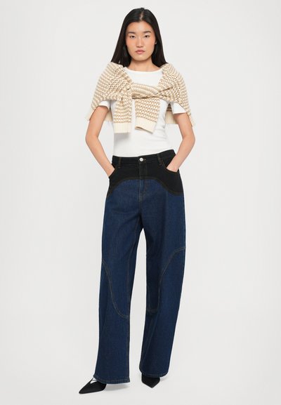 Laiad tumesinised teksad musta vööga ja kaarjate õmblustega; mudel kannab kreemjat kootud salli, mis on juhuslikult valge köitega üle visatud.