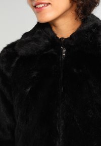 Giacca in pelliccia ecologica nera con chiusura a zip, collo alto e superficie morbida e testurizzata, che presenta un design elegante e voluminoso.