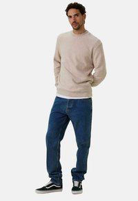 Pull en polaire beige à col rond associé à un jean en denim bleu. Le modèle porte des baskets noir et blanc, debout devant un fond uni.