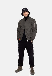 Grüne Bomberjacke mit Reißverschluss und Taschenakzenten, schwarzer Rollkragenpullover, schwarze Cargo-Hosen, grüne Stiefel und ein schwarzer Bucket Hat.