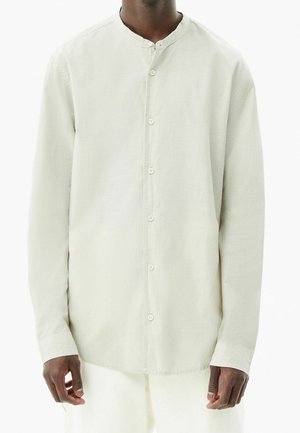 Uomo che indossa una camicia a maniche lunghe color avorio con collo alla mandarina e bottoni, in piedi davanti a uno sfondo bianco neutro.