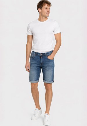Junger Mann steht mit einer Hand in der Tasche, trägt ein schlichtes weißes T-Shirt, hochgekrempelte blaue Jeansshorts und weiße Sneaker.