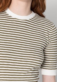 Primer plano de una persona que lleva una camisa de manga corta a rayas horizontales en blanco y verde oliva, con un cuello blanco.