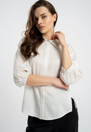 Kobieta o długich brązowych włosach, ubrana w białą koszulę z guzikami i marszczonymi rękawami oraz czarne spodnie, pozująca na tle gładkiego, jasnego tła.