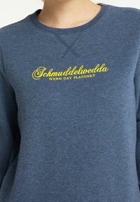 Sudadera azul hecha de un tejido texturizado, que presenta un texto bordado en amarillo que dice "Schmuddelwedda WENN DAT PLADDERT."