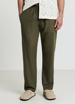 Pantaloni verdi oliva a vestibilità rilassata con vita elasticizzata, dotati di due tasche frontali e texture liscia. Abbinati a scarpe beige slip-on.