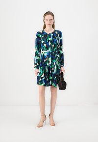 O vestido apresenta um padrão abstrato multicolorido em azul, verde e preto, com mangas compridas, tecido plissado e um cinto fino branco. Uma pequena handbag complementa o visual.