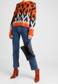 Pull en maille à motif léopard orange et noir, avec une coupe courte. Porté avec un jean bleu taille haute et des bottines rouges à talons, tenant un sac noir.