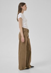 Pantalones de pierna ancha marrones con una tela texturizada, de tiro alto y con pliegues en la parte delantera, combinados con una camiseta ajustada blanca y zapatos negros de punta.