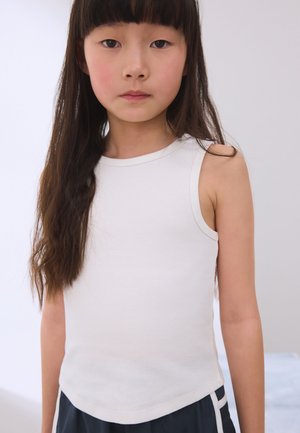 Jong meisje met lang donker haar, gekleed in een wit mouwloos topje en donkere korte broek, staand voor een effen lichte achtergrond.