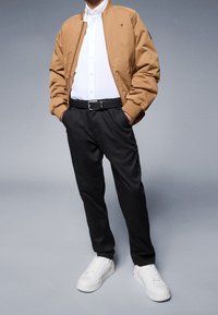 Uomo vestito con una giacca bomber beige, camicia bianca con bottoni, pantaloni neri, cintura nera e sneakers bianche, in piedi con le mani in tasca.