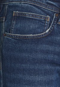 Jeans in denim blu scuro presentano una tasca posteriore con un bordo sfilacciato, cuciture gialle a contrasto e una superficie liscia tipica del tessuto denim.
