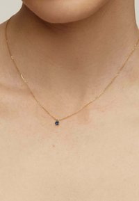 Collar de cadena de oro con un pequeño colgante de gema azul redonda, posicionado delicadamente sobre piel desnuda. La cadena tiene una textura retorcida.