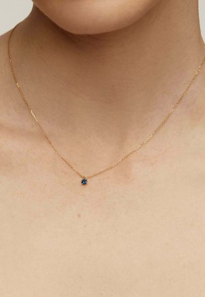 Collar de cadena de oro con un pequeño colgante de gema azul redonda, posicionado delicadamente sobre piel desnuda. La cadena tiene una textura retorcida.