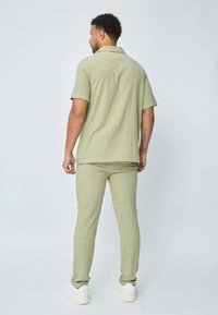 Chemise légère à manches courtes vert olive associée à un pantalon slim assorti. Le tissu présente une texture subtile. Des baskets blanches complètent la tenue.