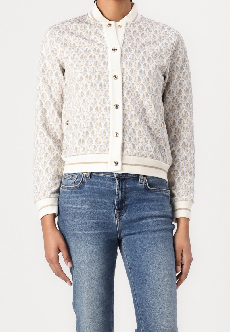 Personne portant une veste beige et blanche à motifs, boutonnée avec des poches, associée à un jean bleu en denim.