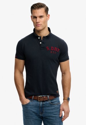 Man draagt een marineblauw poloshirt met de tekst "S. DRY N.Y.C", blauwe spijkerbroek, bruine riem en polshorloge, staand met handen in broekzakken.