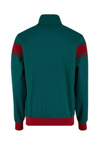 Jachetă de sport de culoare verde închis, cu guler înalt. Prezintă dungi roșii pe umeri și manșete și tiv ribbate roșii. Textură netedă, croială sport.