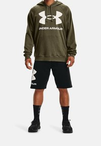 Homme debout portant un hoodie Under Armour vert olive, un short noir avec logo, des chaussettes de sport noires et des chaussures de sport noires, isolé sur fond blanc.