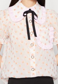 Blouse blanche transparente avec imprimé floral orange et rose, col et poche en vichy rose, nœud en ruban noir et fermetures à boutons dorés.