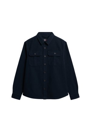 Granatowa koszula zapinana na guziki z grubego materiału; posiada dwie kieszenie na piersi z klapami i długie rękawy. Kołnierz oraz prosty krój na dole.
