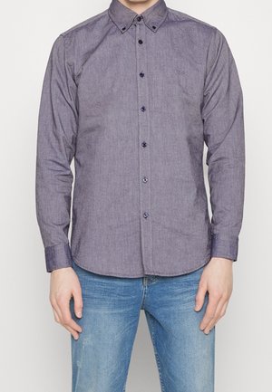 Mann trägt ein dunkelgraues, langärmliges Hemd mit Knöpfen und spitzem Kragen sowie eine blaue Jeans, steht vor einem schlichten Hintergrund.