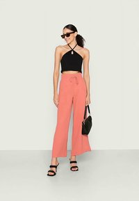 Mulher vestindo um top cortado preto, calças largas corais, sandálias pretas, óculos de sol, segurando uma bolsa preta, de pé contra um fundo claro simples.