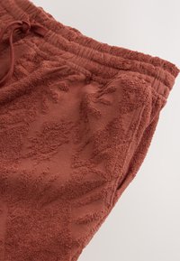 Pantaloni jogger in terracotta con superficie strutturata caratterizzata da un motivo floreale, vita elasticizzata e coulisse per una vestibilità regolabile. Tessuto morbido.