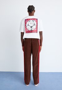 White t-shirt with a graphic print on the back, paired with brown corduroy pants featuring red side stripes. Smooth textures and relaxed fit.