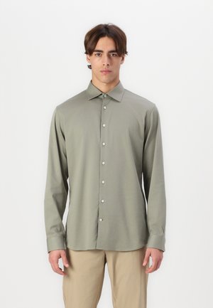 Chemise à manches longues avec boutons, de couleur olive discrète, en tissu lisse, avec un col pointu et des boutons clairs, portée avec un pantalon beige.