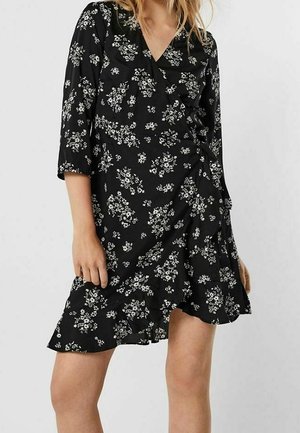 Frau trägt ein schwarzes Wickelkleid mit weißen Blumenmustern und dreiviertelÄrmeln, steht vor einem einfarbigen Hintergrund.
