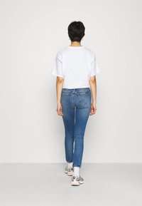 Persona con cabello corto y oscuro, vistiendo una camiseta blanca, unos jeans ajustados azules con bolsillos traseros, calcetines blancos y zapatillas plateadas, de pie contra una pared blanca.