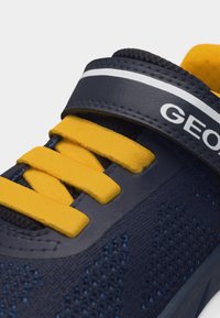 Σκούρο μπλε αθλητικό παπούτσι με κίτρινες κορδόνια, υφασμάτινη επιφάνεια με σχέδιο πλέξης και σκούρο μπλε λουράκι με την ένδειξη "GEOX" και λευκή ρίγα.