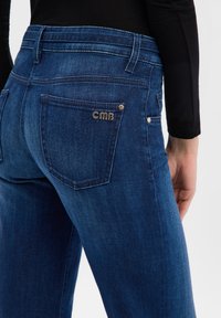 Jean en denim bleu foncé avec une coupe ajustée, doté de poches arrière, de ferrures dorées et d'un petit détail de logo sur la poche droite.
