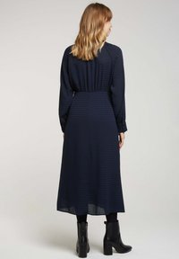 Robe longue à manches longues de couleur bleu marine avec un motif texturé, taille froncée et une silhouette fluide, associée à des bottines noires.